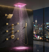 A luxury matte gold shower system installed on a dark marble wall, featuring a square rain shower head with pink illuminated water spray, a handheld wand, and multiple control valves. A large window on the right shows a glowing city skyline at night.