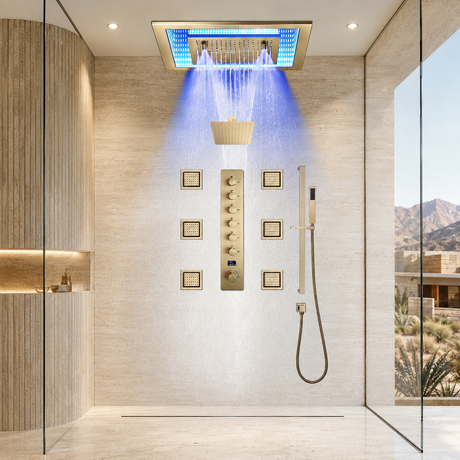 A luxury modern shower system installed in a beige stone-walled bathroom. The system features a rectangular rain shower head with blue LED lighting, a central vertical control panel with gold-toned knobs, a handheld wand with a hose, and a basket-style filter.