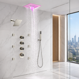 A modern wall-mounted rain shower system with a square pink LED-lit overhead shower head, multiple control valves, a handheld wand, and visible water spray, installed in a marble-look bathroom with a glass door and city view.