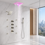 A modern wall-mounted rain shower system with a square pink LED-lit overhead shower head, multiple control valves, a handheld wand, and visible water spray, installed in a marble-look bathroom with a glass door and city view.